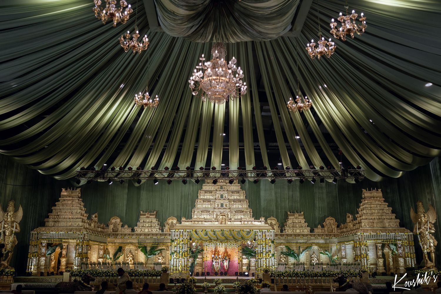 Elegant draped fabric ceiling with crystal chandeliers hanging above an ornate Indian wedding stage decoration.