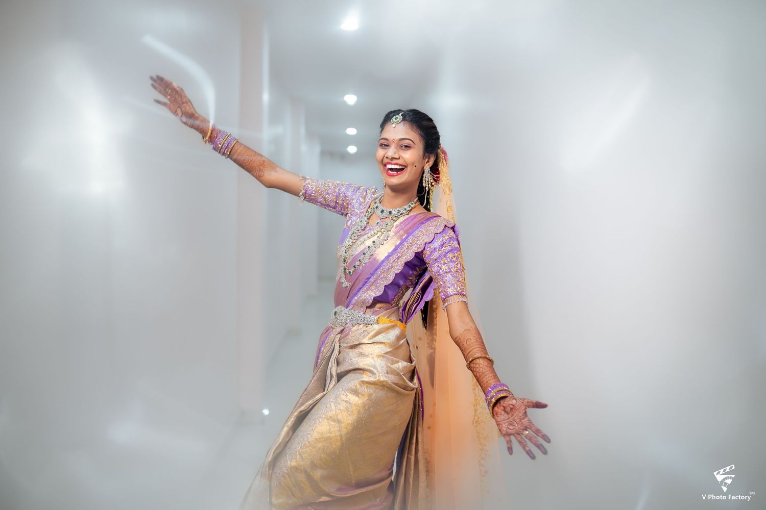 A dancer in a gold and purple silk saree strikes an expressive dance pose against a misty white backdrop.