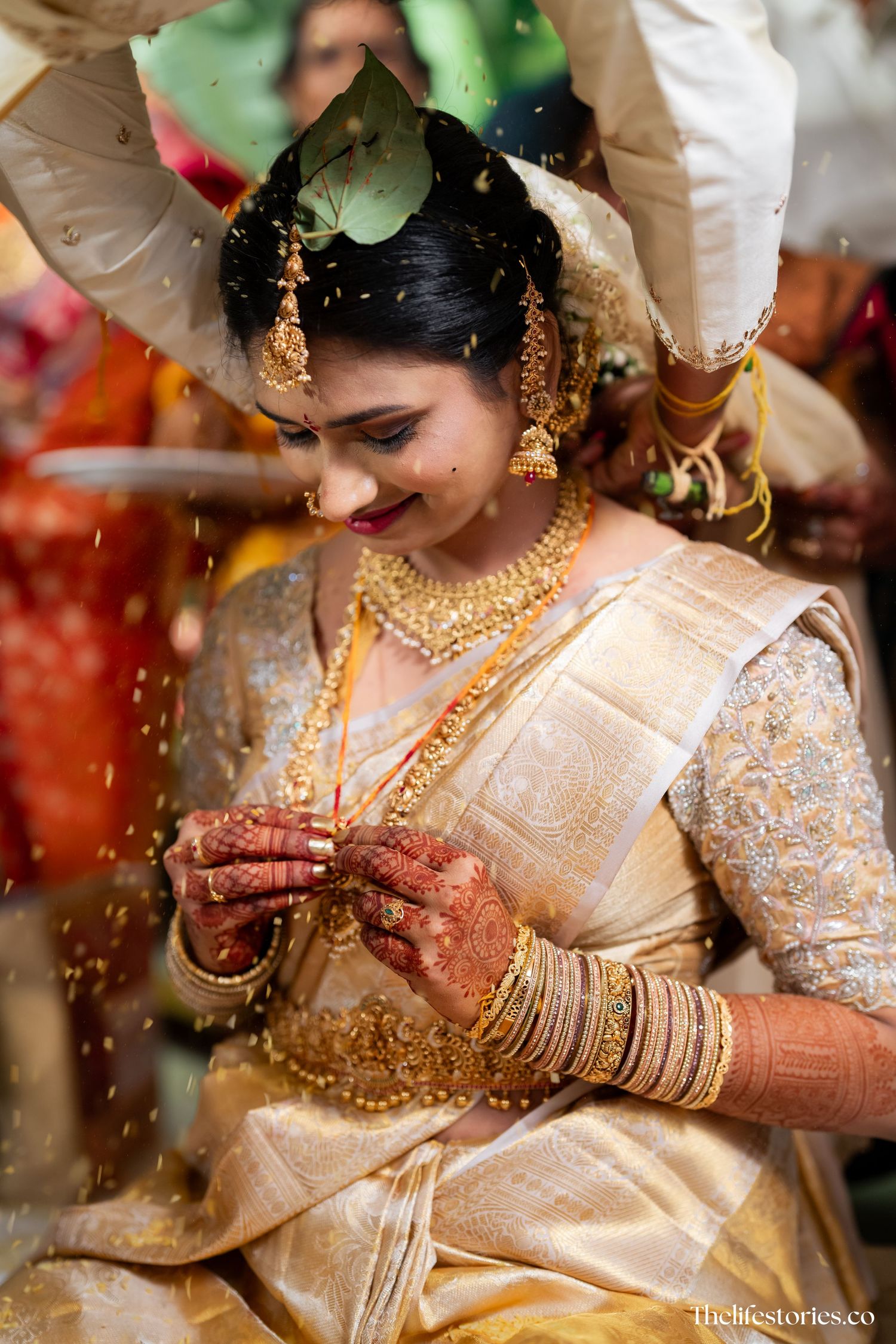 Traditional South Indian bride wearing elegant cream silk saree with gold jewelry and intricate henna designs on her hands.