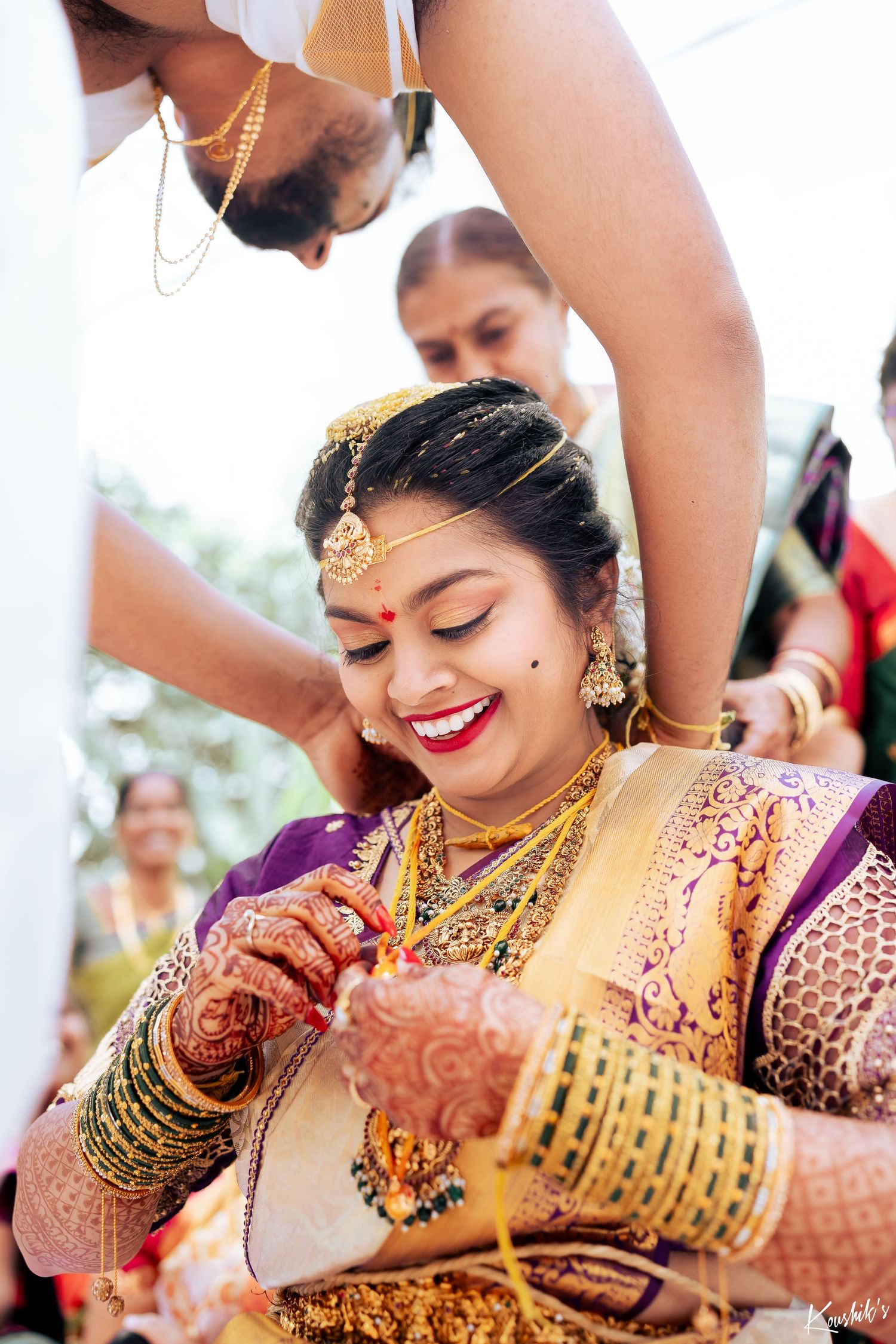 A South Indian bride wearing a purple and gold silk saree smiles while getting adorned with traditional wedding jewelry.