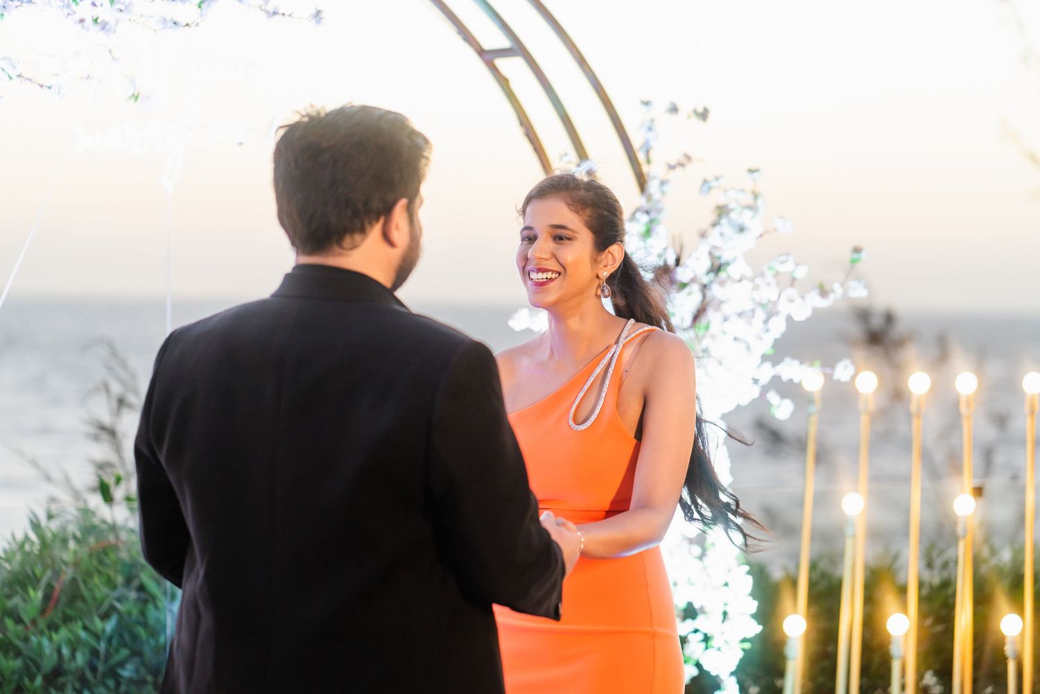 Two people share a joyful moment during a sunset proposal on a terrace with decorative lights and floral arrangements.