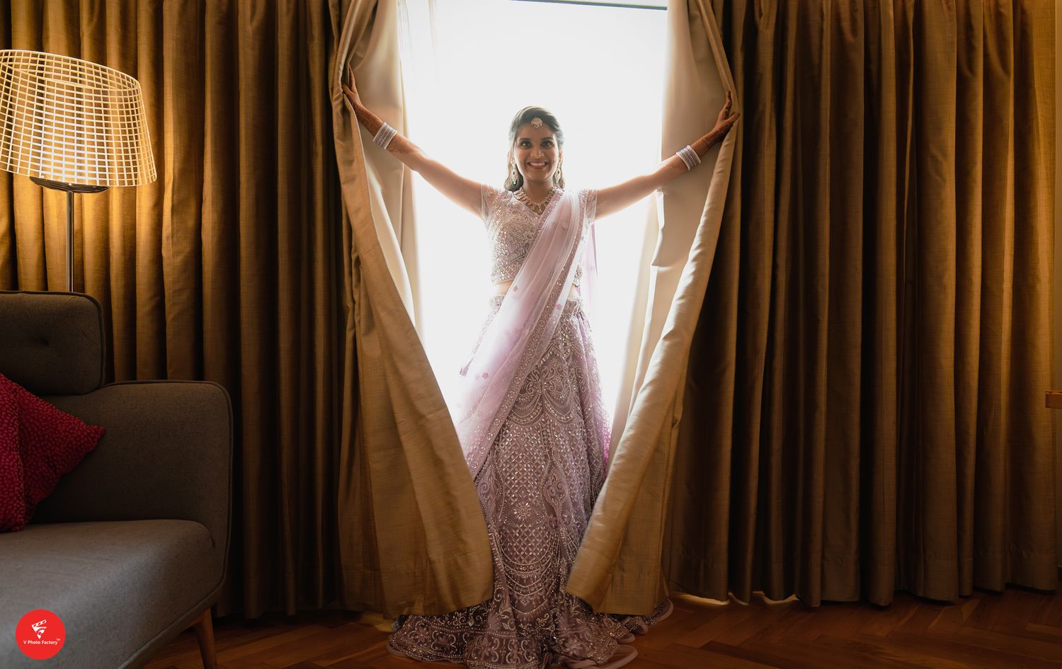A figure in a flowing white wedding gown stands gracefully between parted gold curtains with soft window light behind.