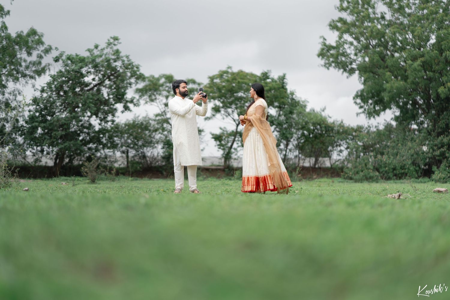 A couple in traditional wedding attire stands together on a grassy hill surrounded by trees.