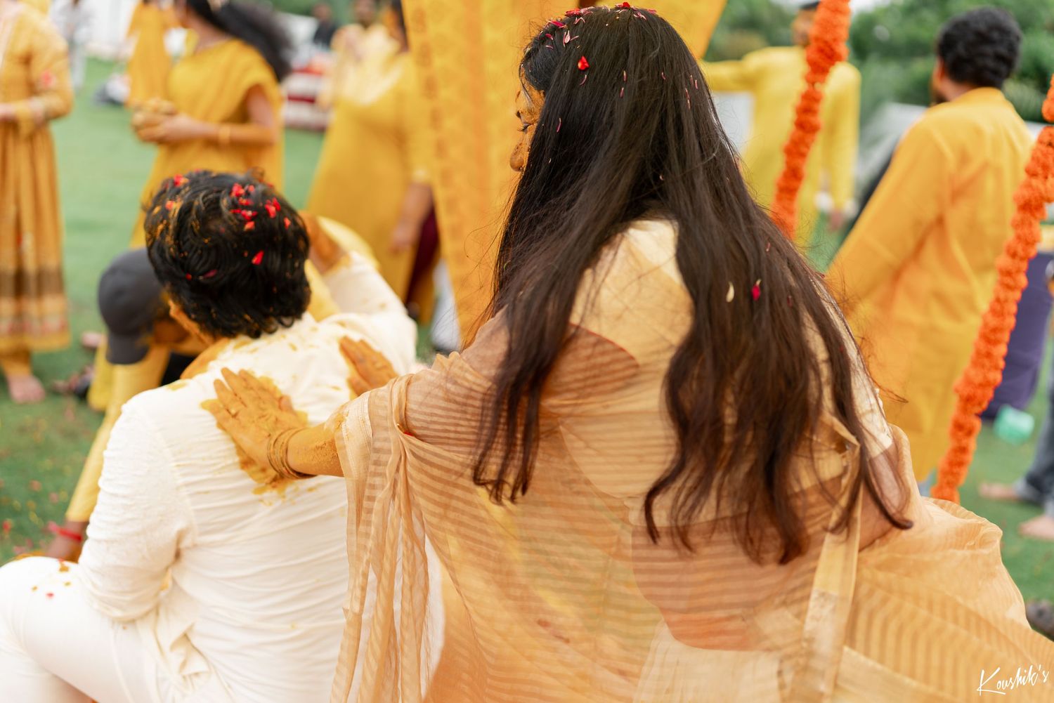 People in flowing yellow and white outfits gather outdoors on grass with long-haired figures visible from behind.