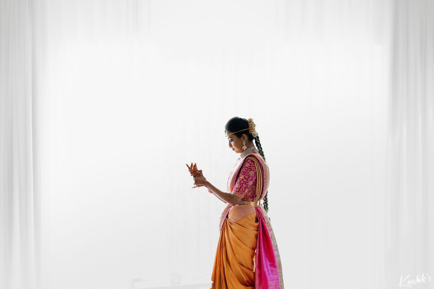 A person wearing a traditional Indian saree in orange and pink looks at their mobile phone in front of a white wall.