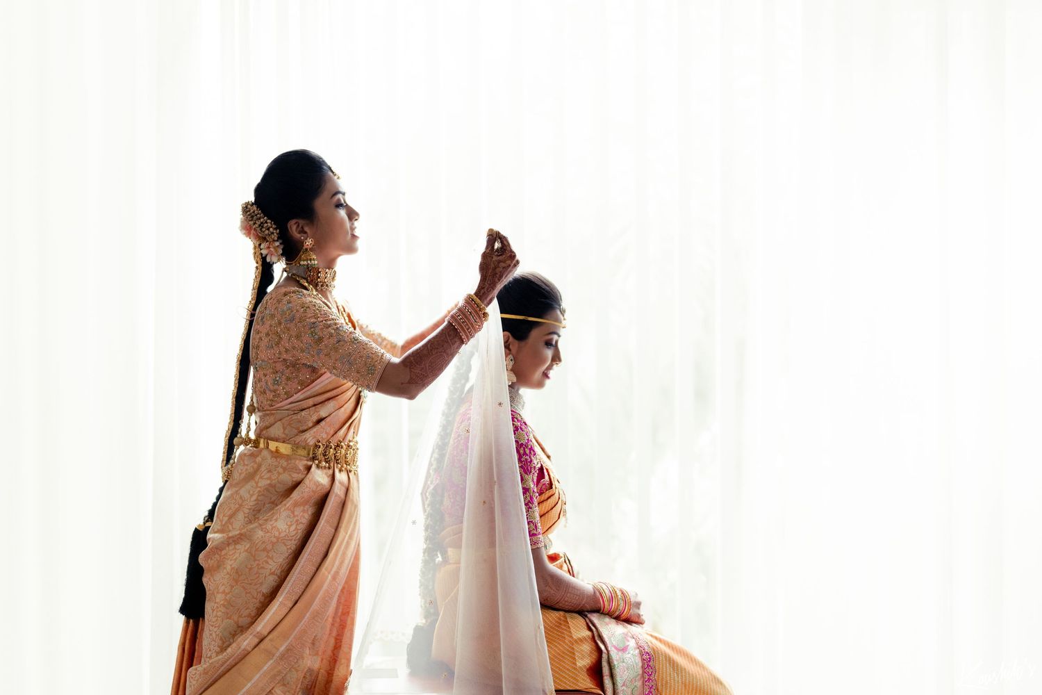 A bride getting fitted with her white wedding veil while wearing a traditional peach and pink embroidered dress.