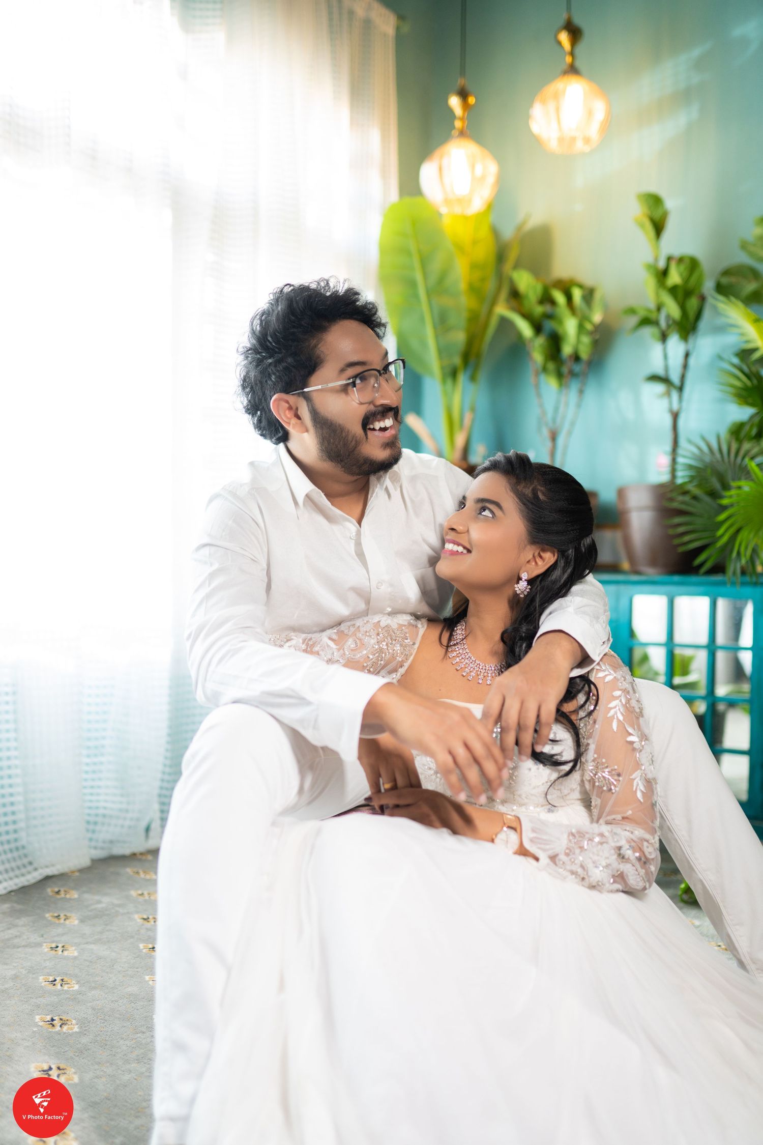 A couple in white traditional outfits share a joyful moment together against a backdrop with hanging lights and plants.