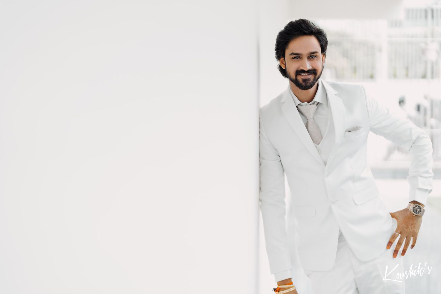 A person wearing a crisp white suit leans against a white wall while smiling at the camera.
