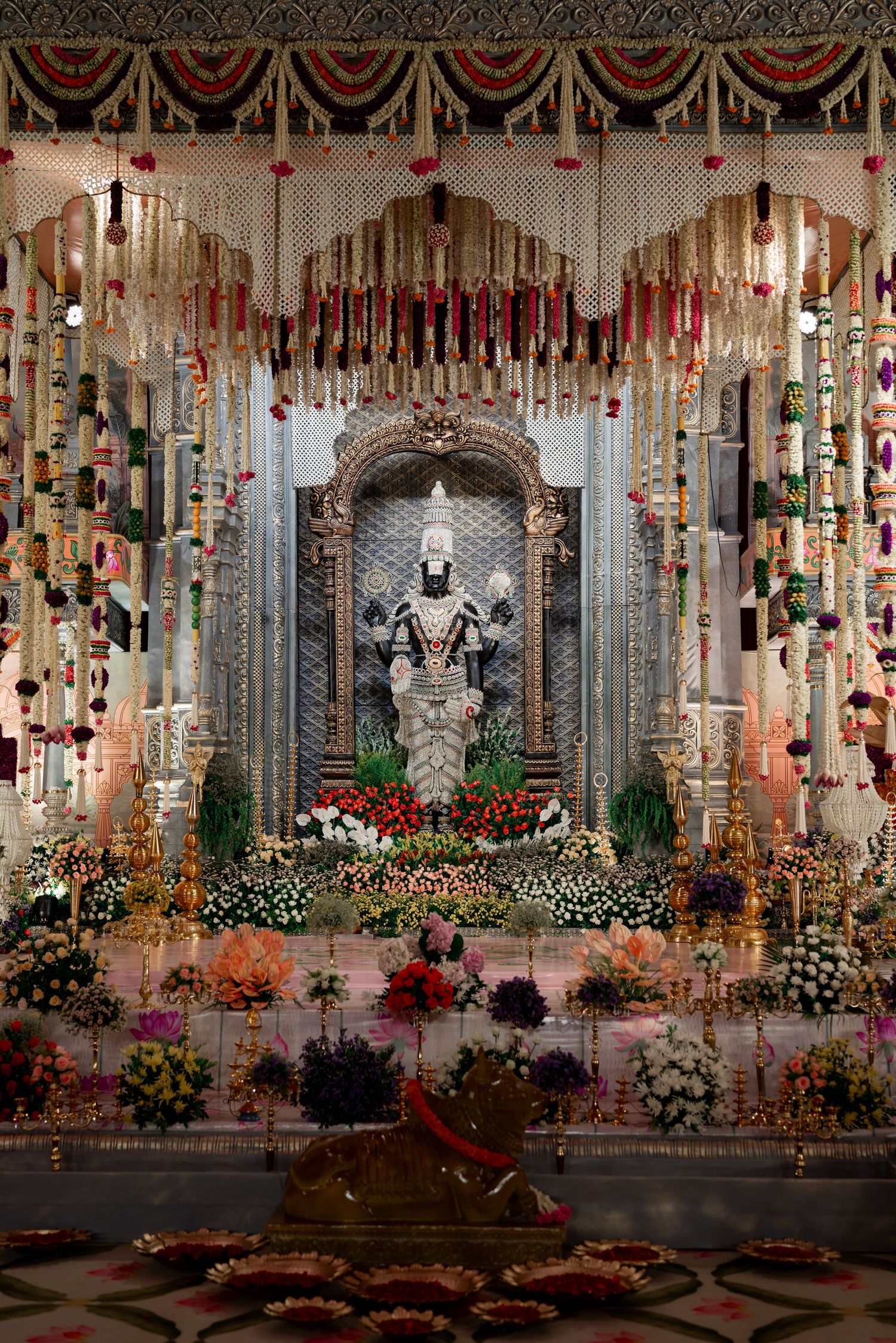 Ornate temple altar decorated with hanging flowers, lights and decorative drapes featuring a shrine with spiritual statue.