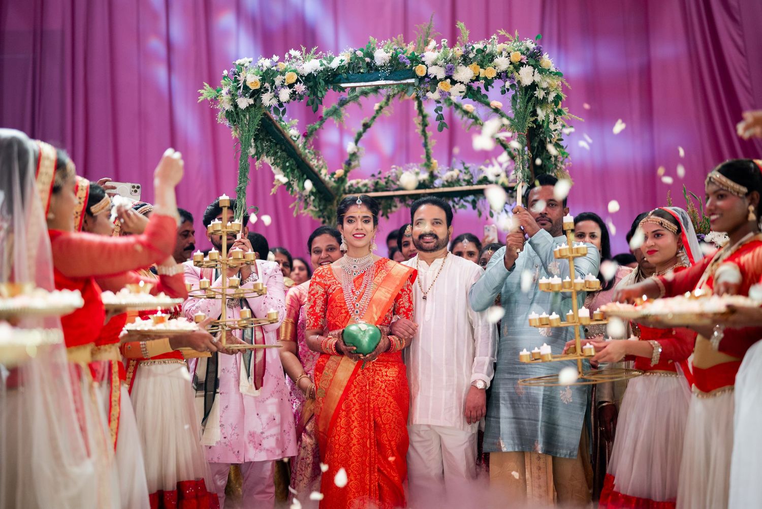 Wedding celebration under floral arch with guests dancing and wearing traditional outfits in pink and red tones.