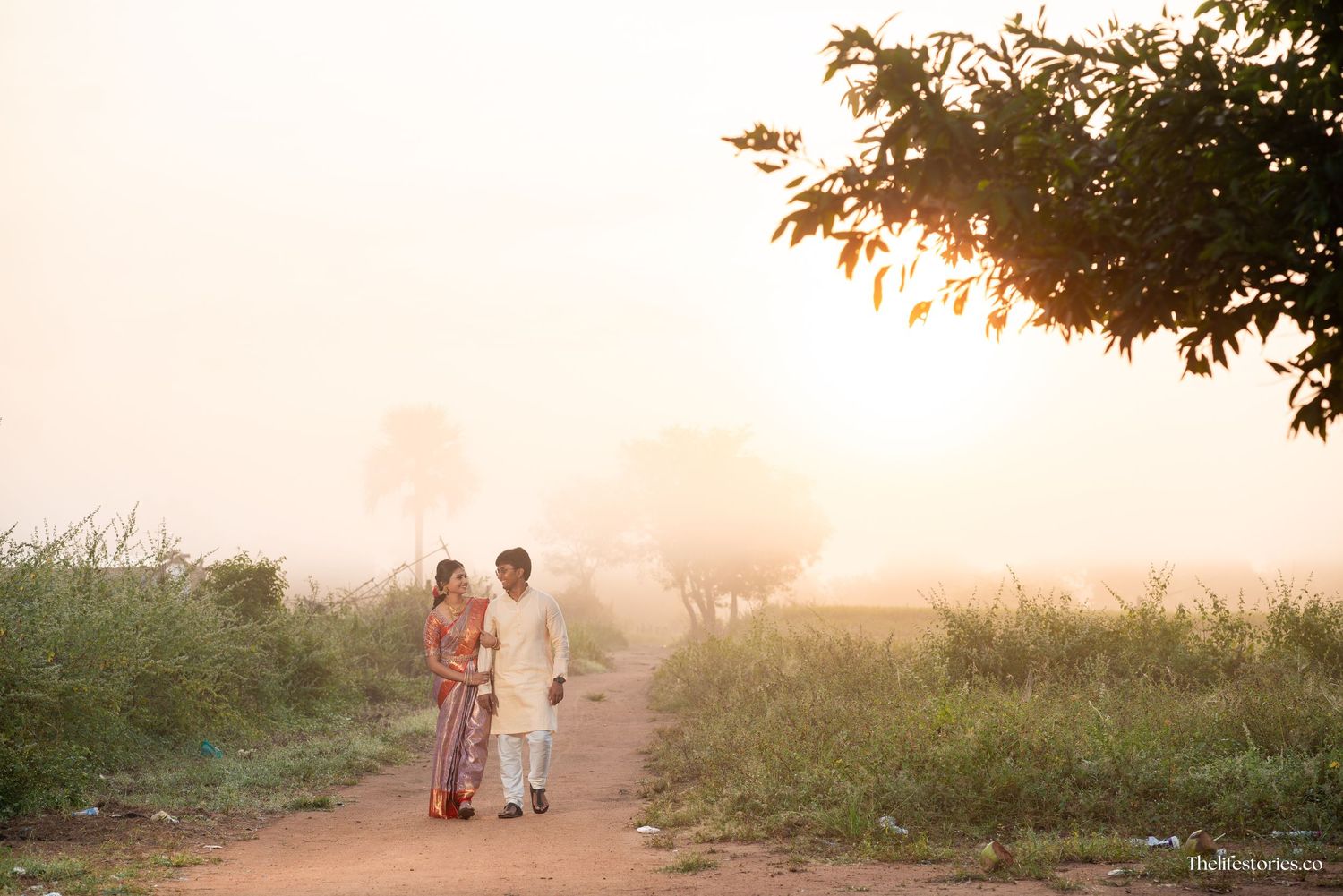 Two people walk down a misty dirt path at sunset with golden light filtering through trees in the background.