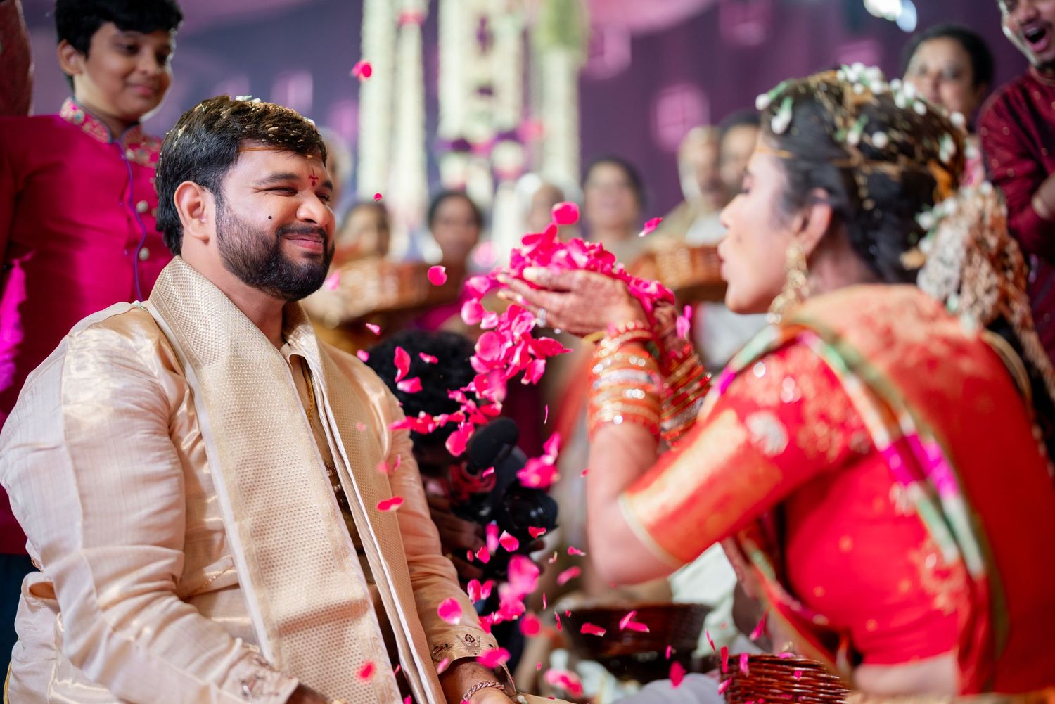A couple exchanges pink flowers during a traditional Indian wedding ceremony with colorful outfits and decor.