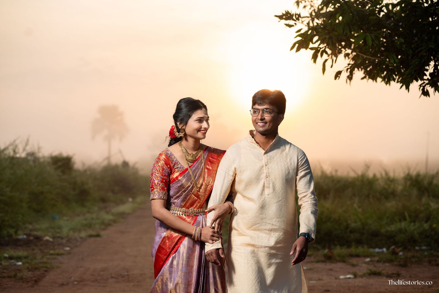 A couple in traditional Indian attire walk together on a rural path during sunset, with trees in the background.