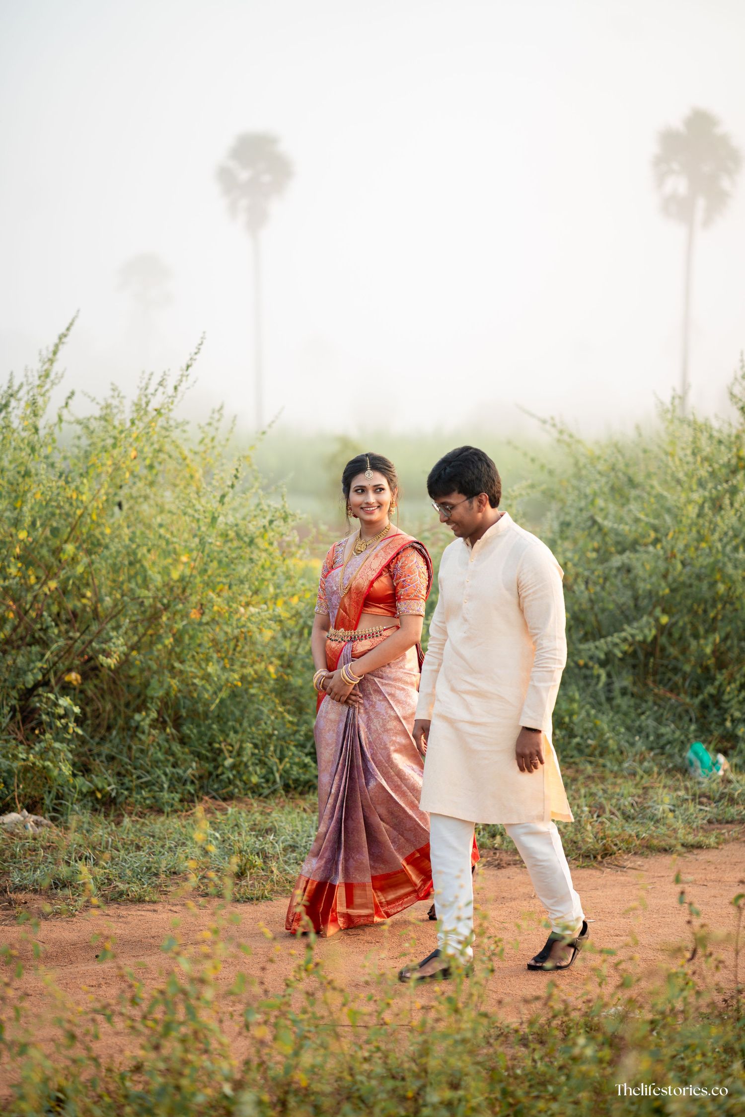 A couple in traditional Indian attire walks together through a rural setting with palm trees in the misty background.