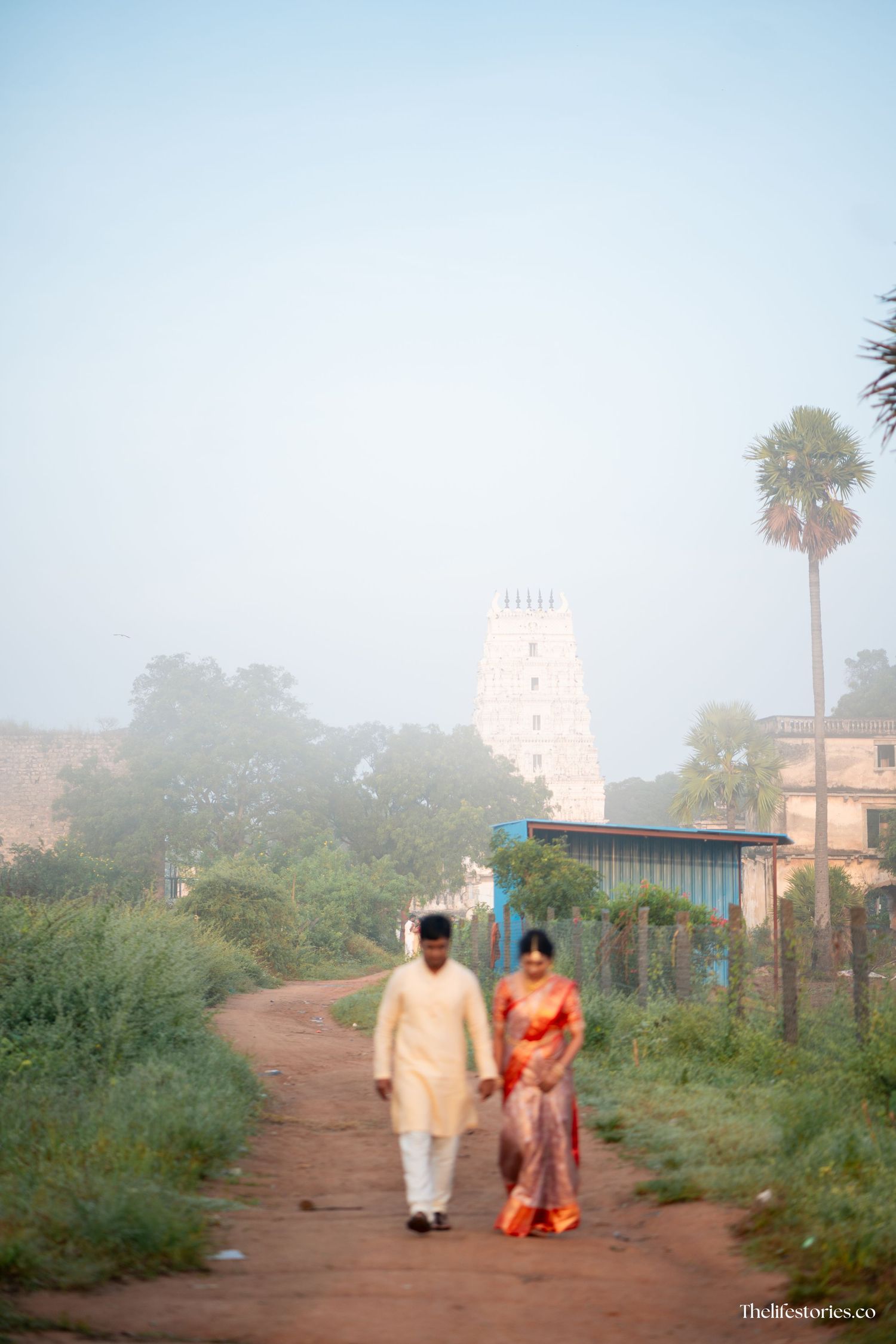 Two figures walk on a dirt path with a white temple tower visible in the misty background.