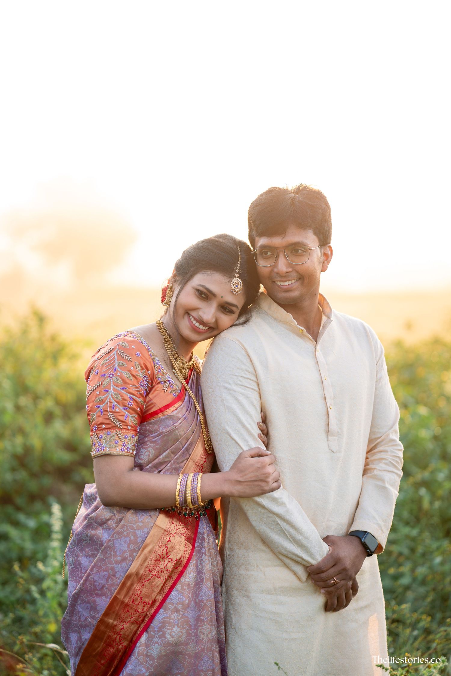 A couple poses in traditional Indian attire during golden hour in a rural setting with natural sunlight.
