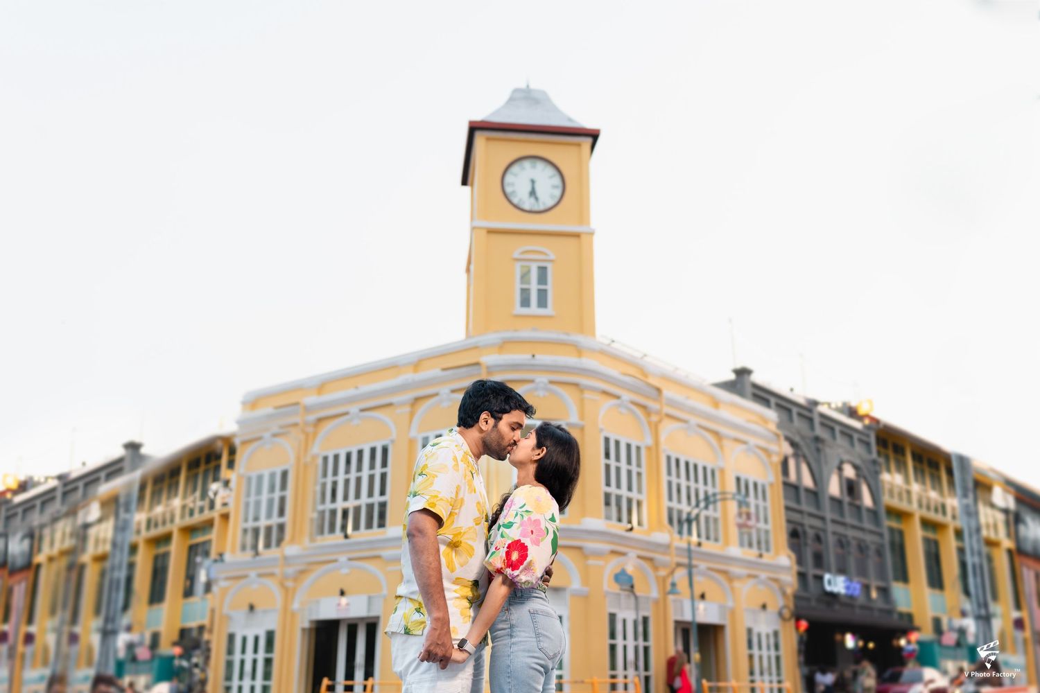 A couple embraces in front of a historic yellow building with a clock tower in Phuket Old Town, Thailand.