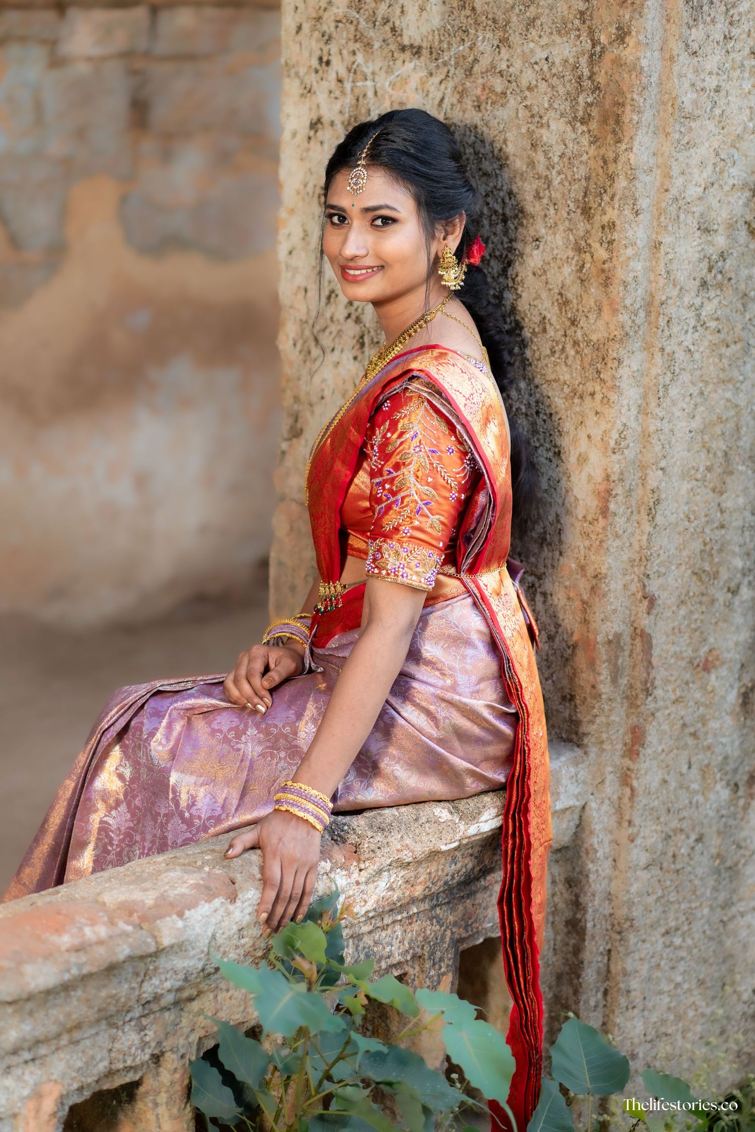 A traditional South Indian silk saree with golden border and red blouse posed elegantly against a rustic stone wall.