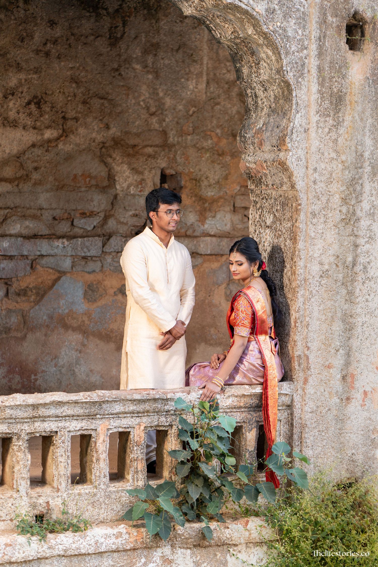 A couple in traditional Indian attire pose in an ancient stone archway with ivy growing on the walls.