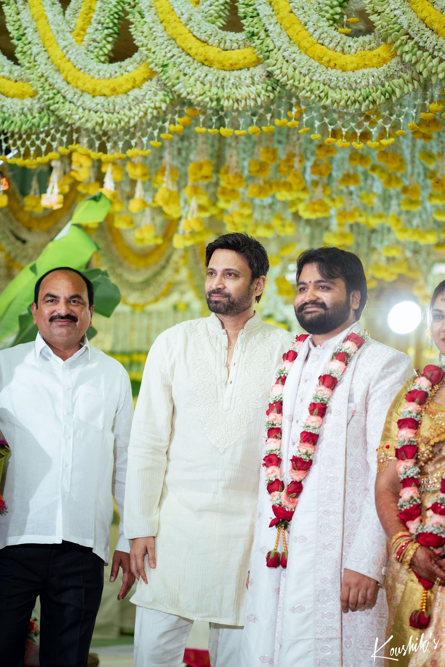 Three men in traditional white Indian attire stand together under decorative yellow and green wedding mandap decorations.