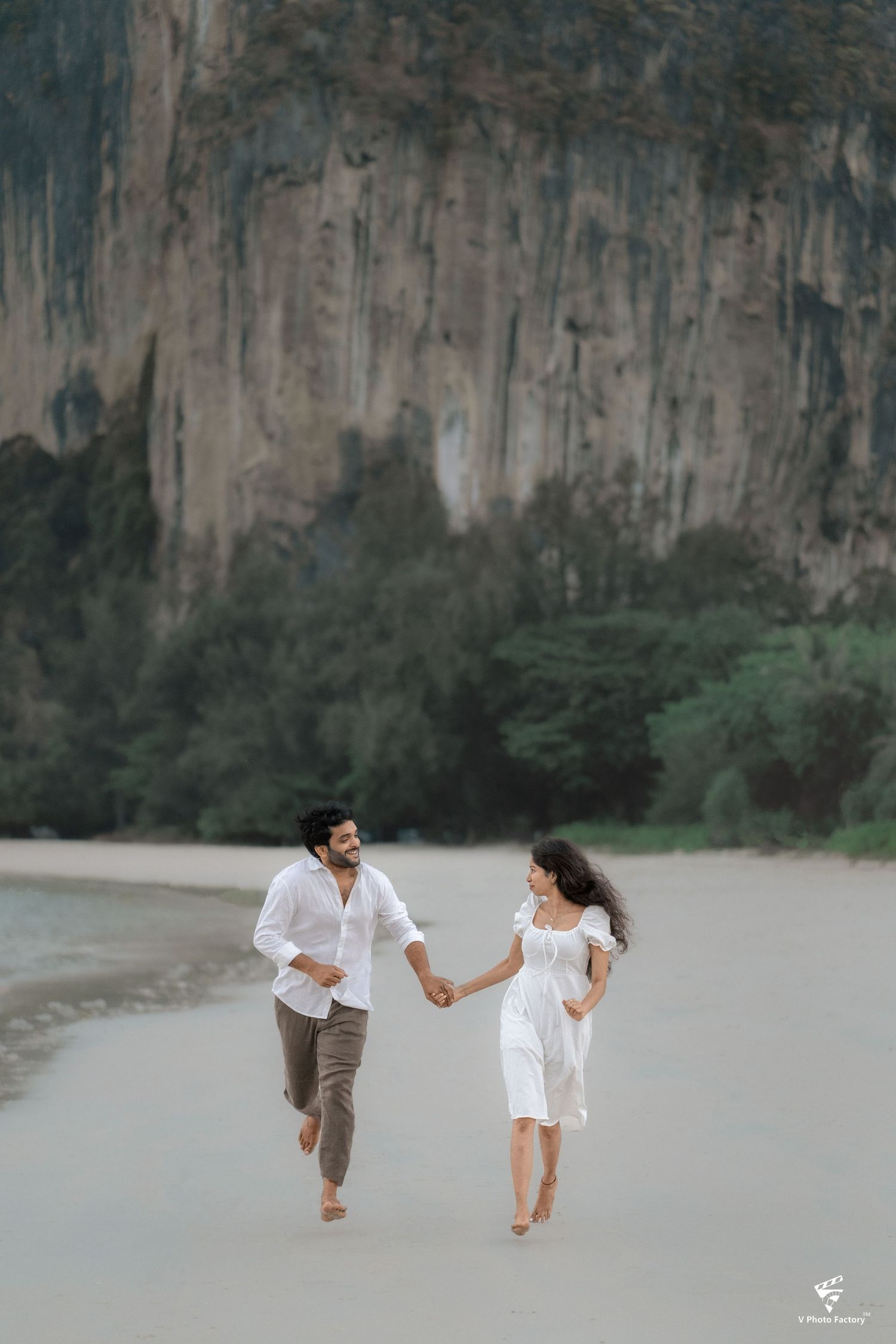 A couple runs barefoot along a beach with dramatic limestone cliffs in the background.