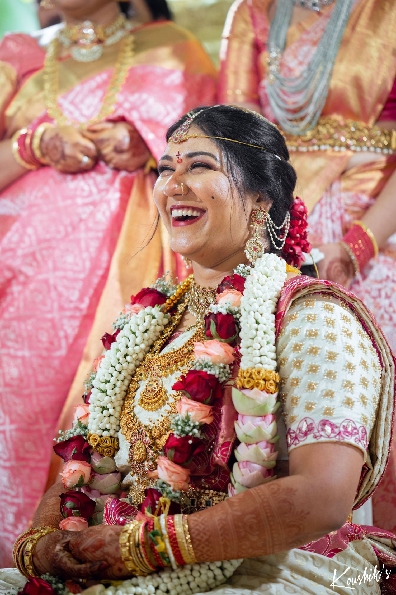 Beautiful South Indian bride in traditional attire wearing floral garlands and gold jewelry laughs joyfully during ceremony.