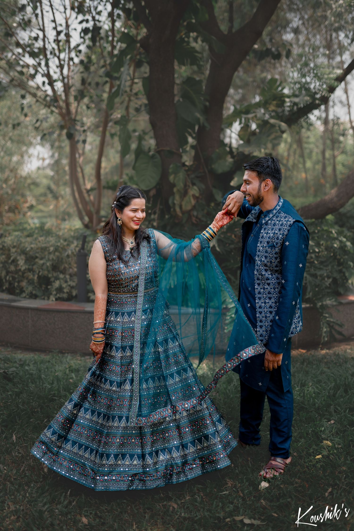 A couple in coordinating blue Indian traditional outfits pose together in a romantic outdoor garden setting.