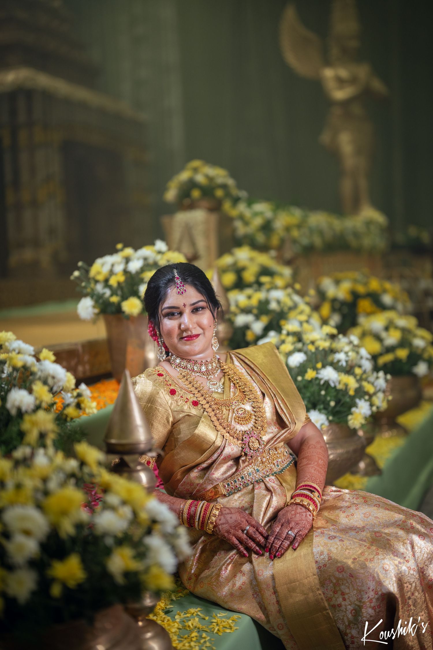 A bride in a traditional gold silk saree and wedding jewelry sits among yellow and white floral decorations at a ceremony.