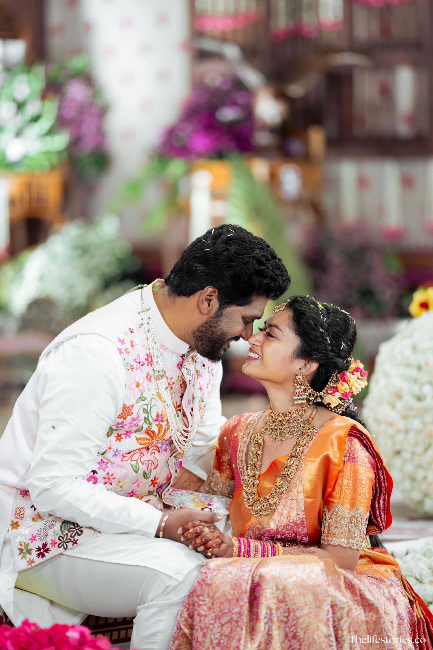 A couple in traditional Indian wedding attire share an intimate moment amidst floral decorations.