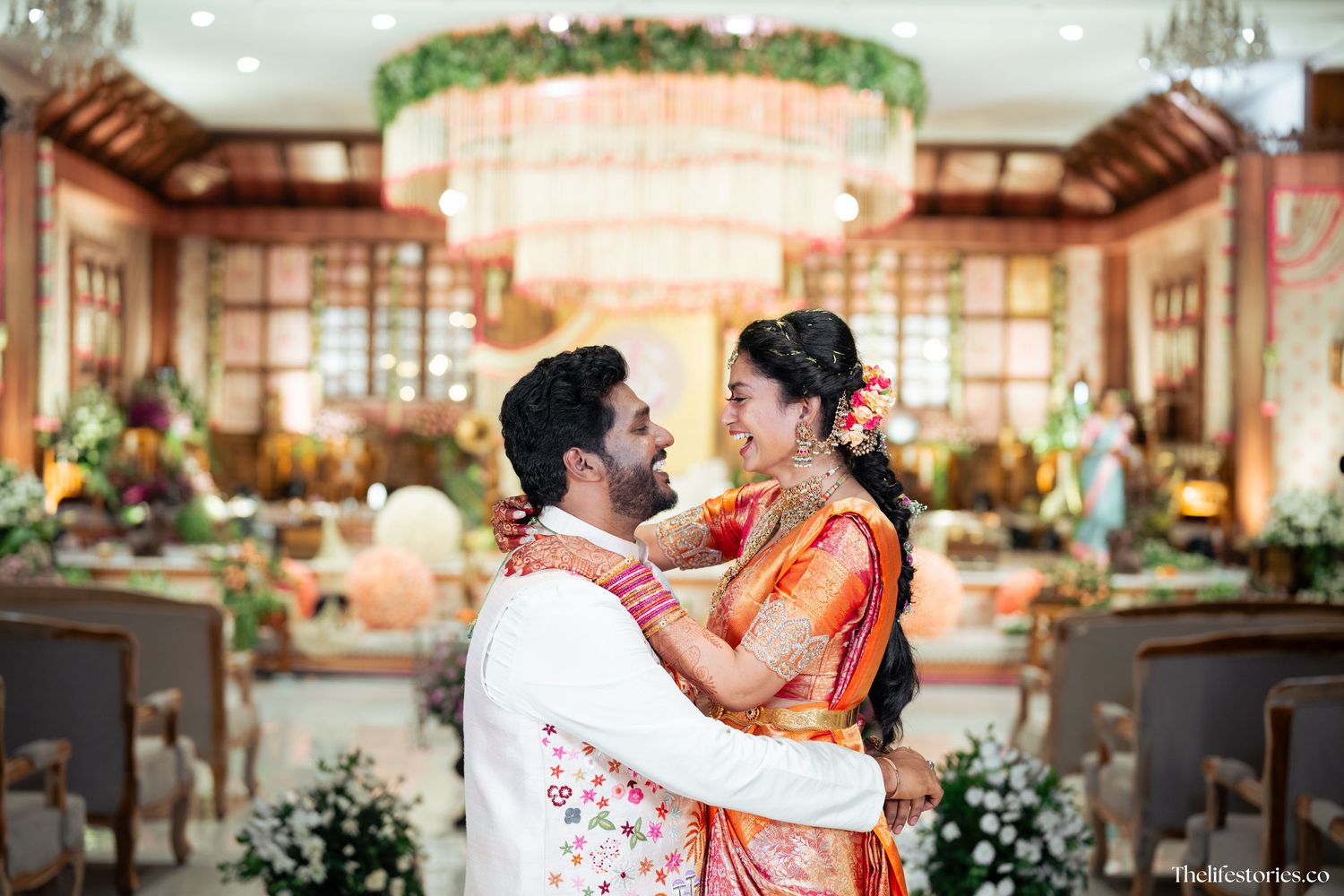 A couple in traditional Indian wedding attire embrace during their celebration in an elegantly decorated indoor venue.
