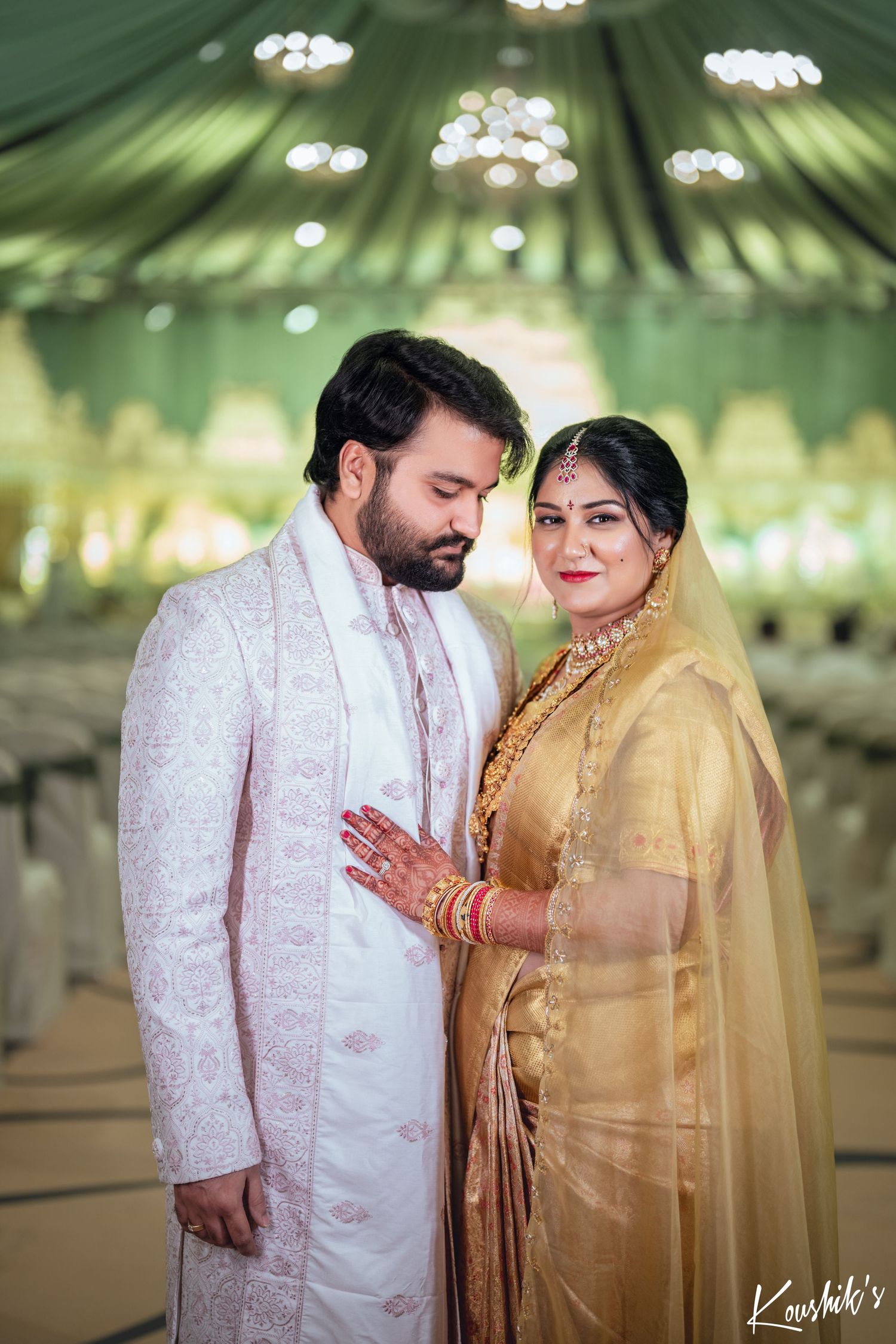 A couple wearing traditional Indian wedding attire pose together under an elegant green-draped ceiling with twinkling lights.