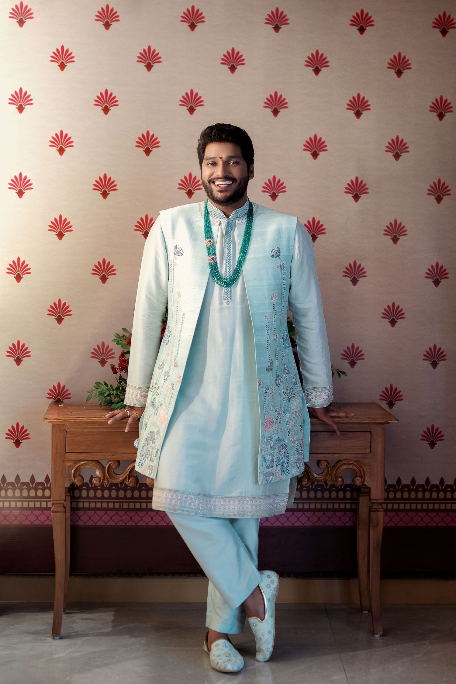 Traditional Indian groom wears light blue and white kurta with embroidered details against floral wallpaper backdrop.