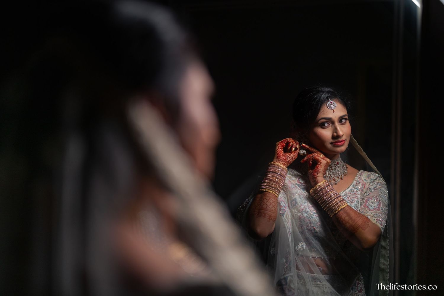 A person wearing traditional Indian bridal attire and jewelry prepares in front of a mirror in dramatic lighting.