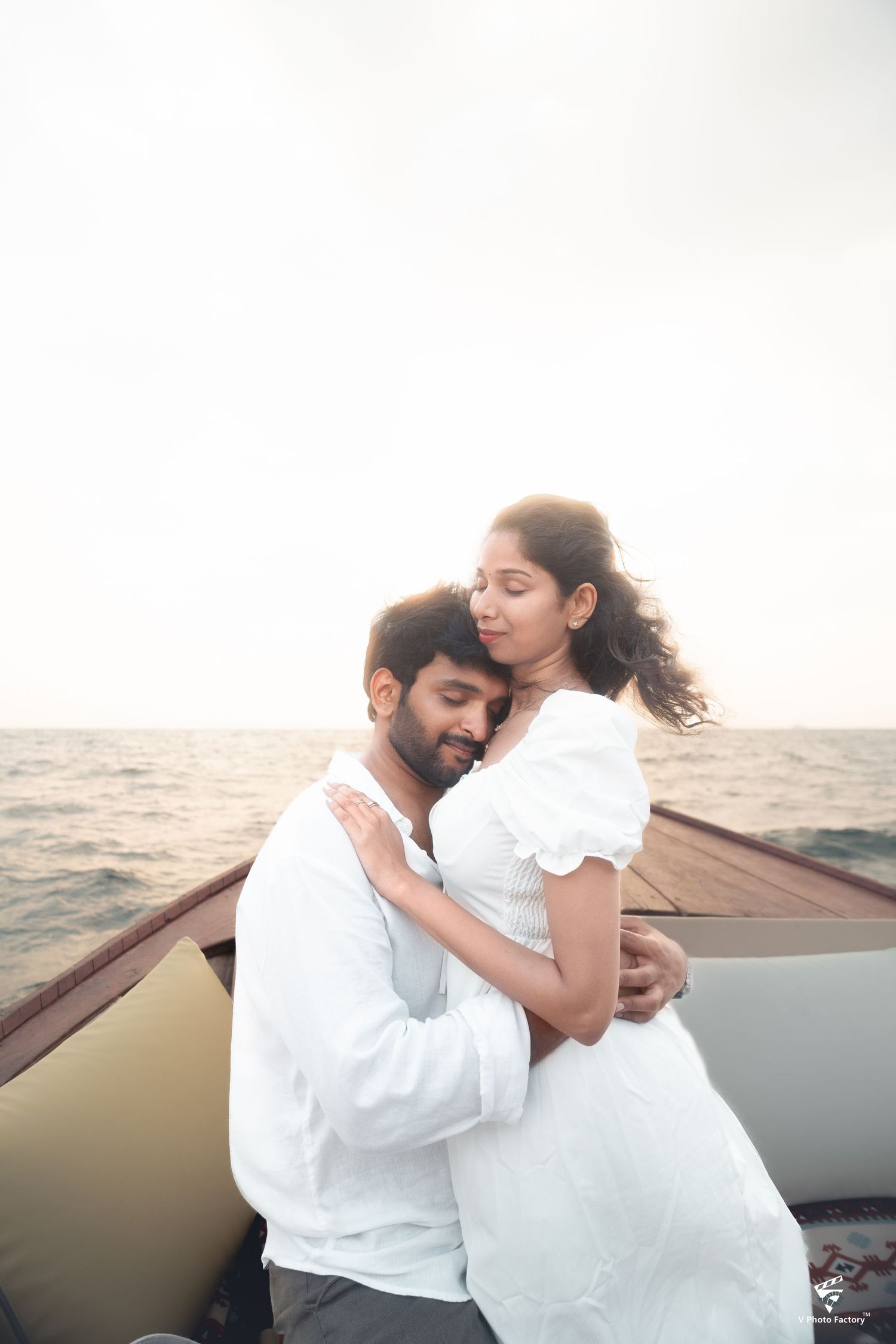 A romantic couple in white outfits embrace while standing on a boat at sea during sunset.