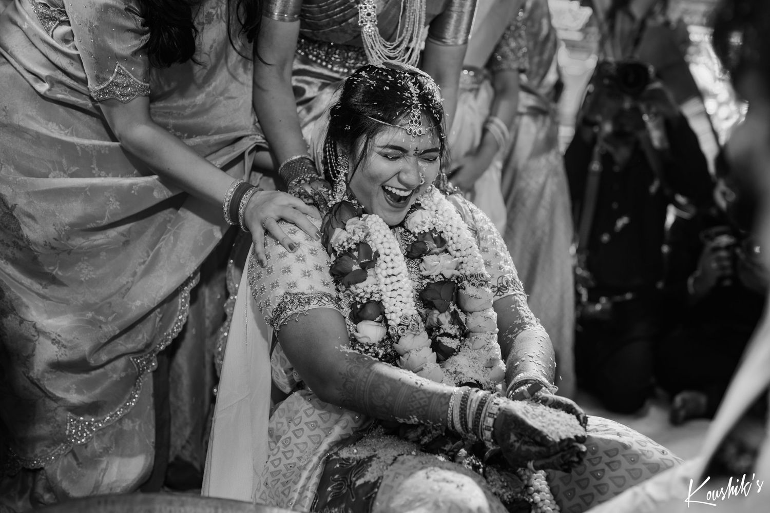 A candid black and white wedding photo showing a joyful seated guest wearing flower garlands during a traditional ceremony.