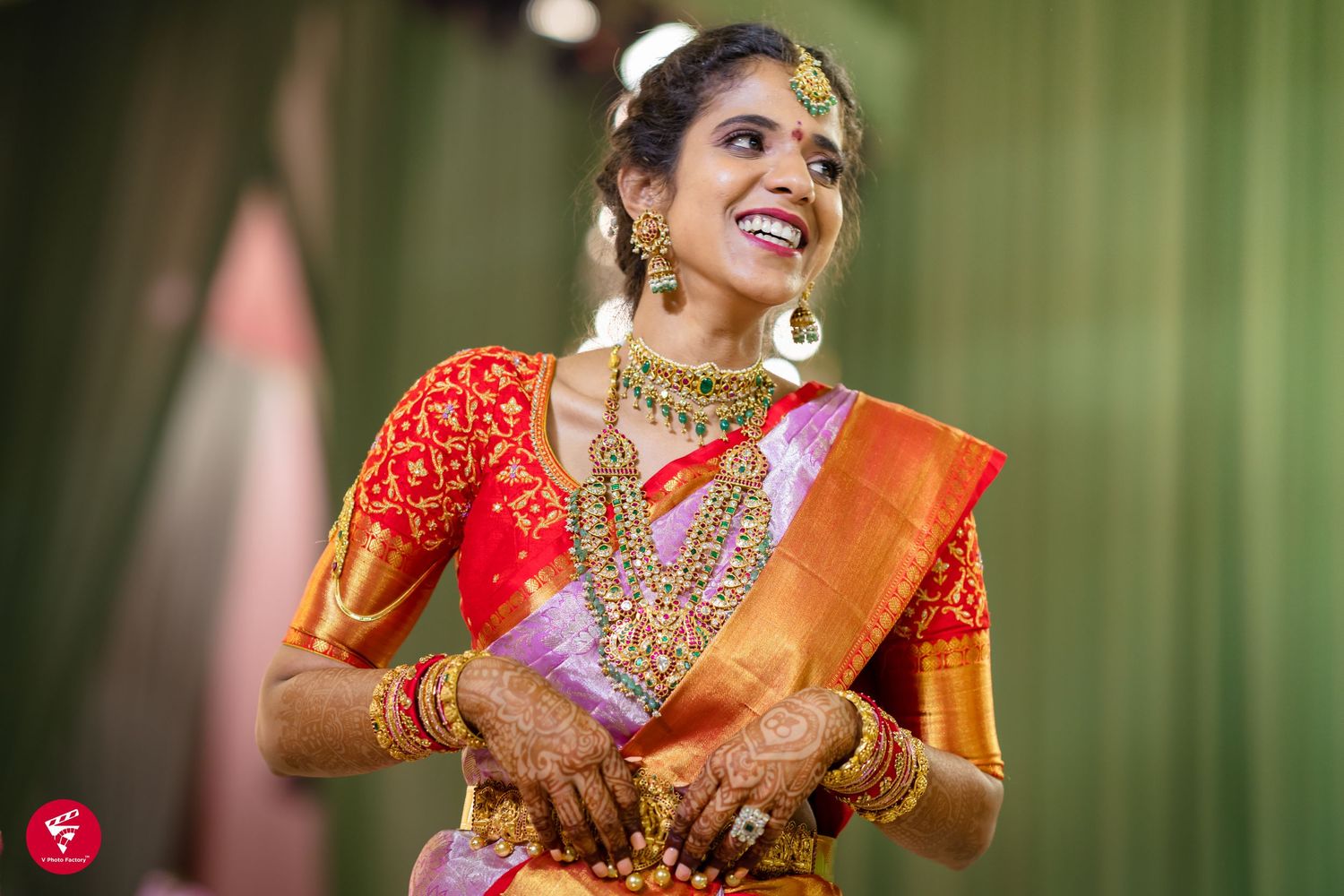 Traditional South Indian bride wearing vibrant red silk saree and gold jewelry smiles during wedding ceremony.