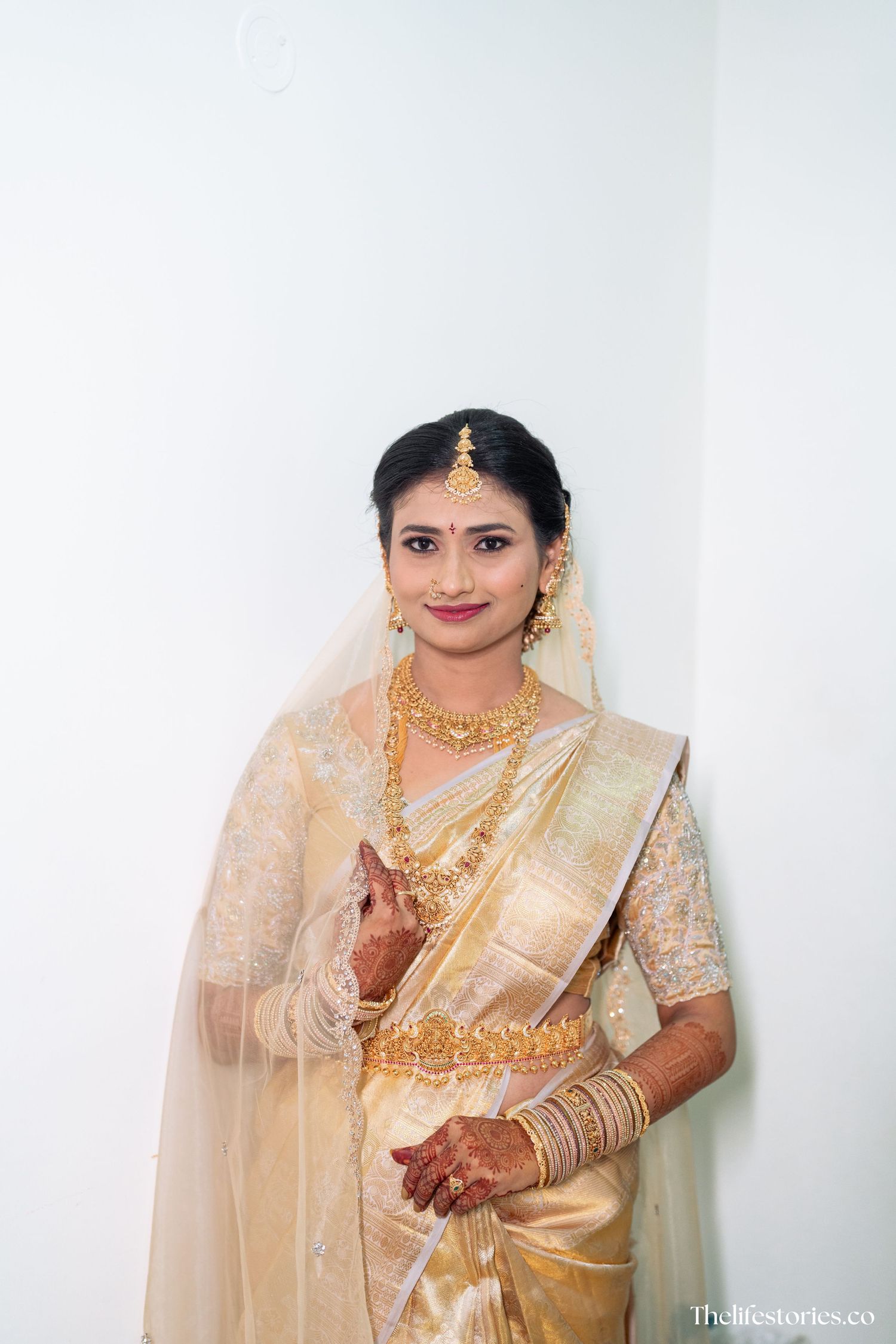 A bride wearing a traditional cream and gold saree with ornate jewelry and henna adorned hands poses elegantly.