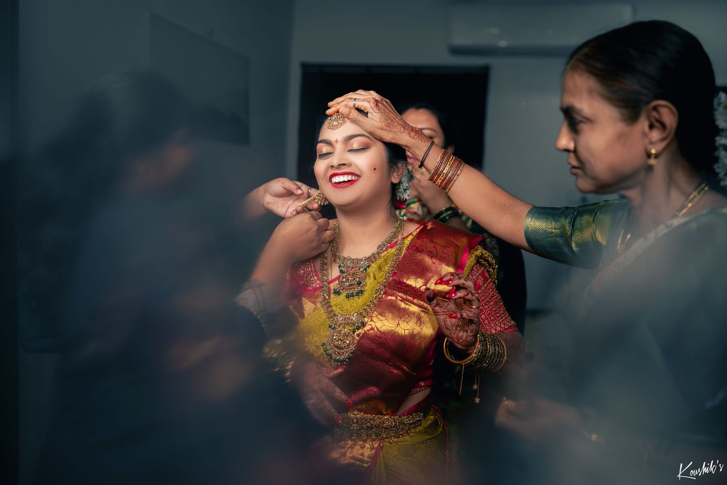 A person in traditional Indian attire and jewelry getting ready while others help with preparations in a moody setting.