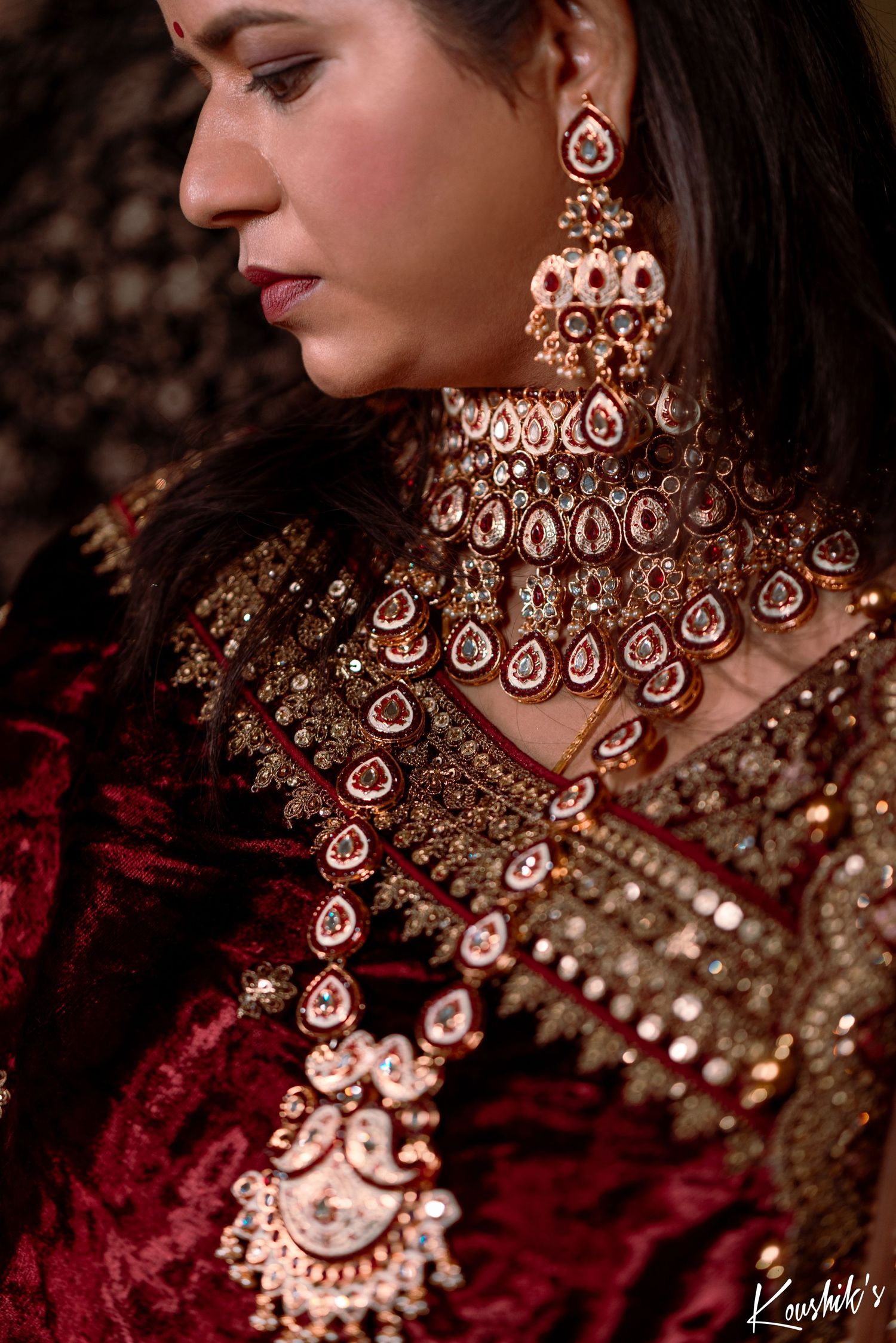 Close up view of ornate Indian bridal jewelry with intricate kundan necklace and chandelier earrings against red velvet attire.