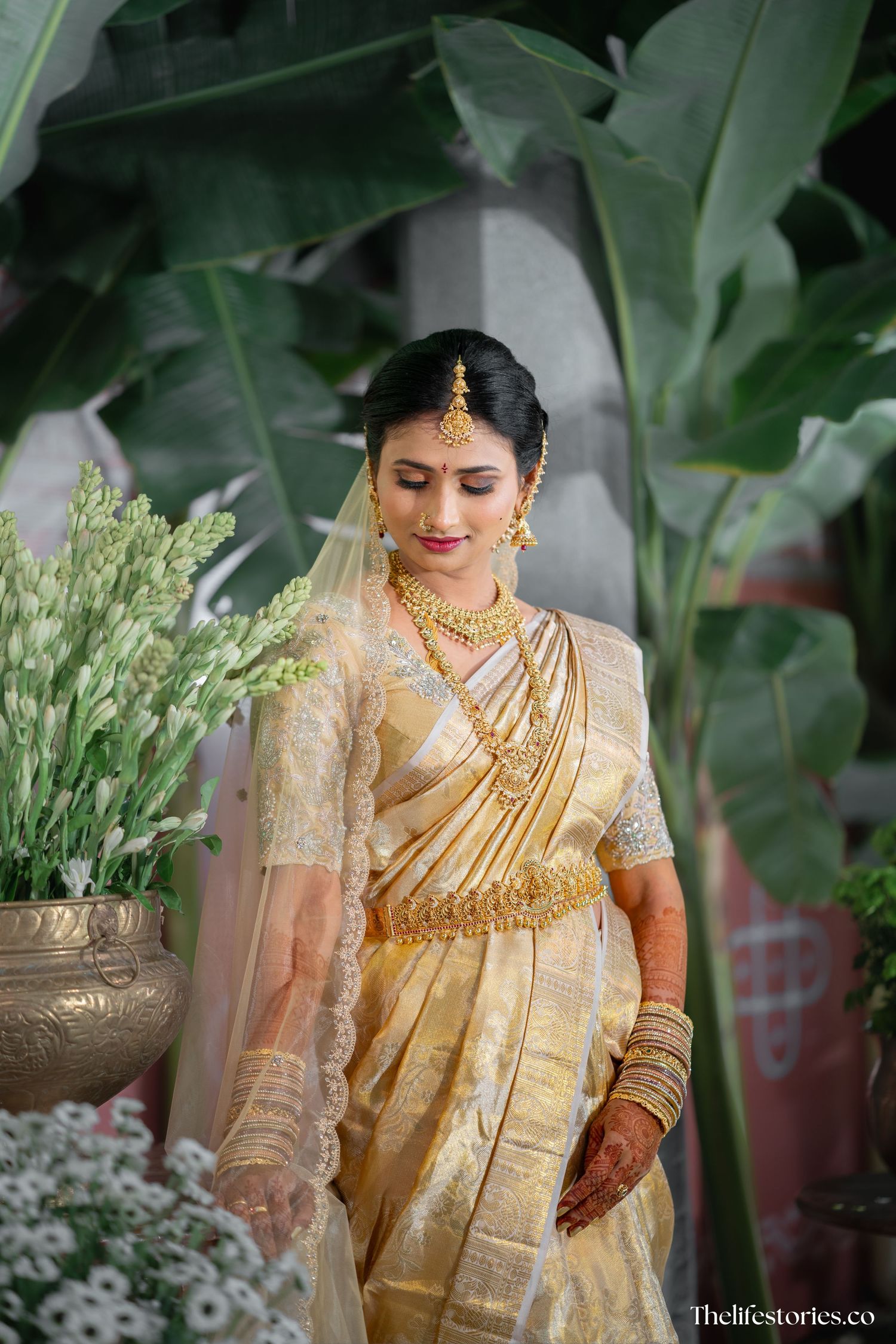 A bride wearing a traditional golden silk saree with jewelry and veil stands elegantly against a green leafy background.