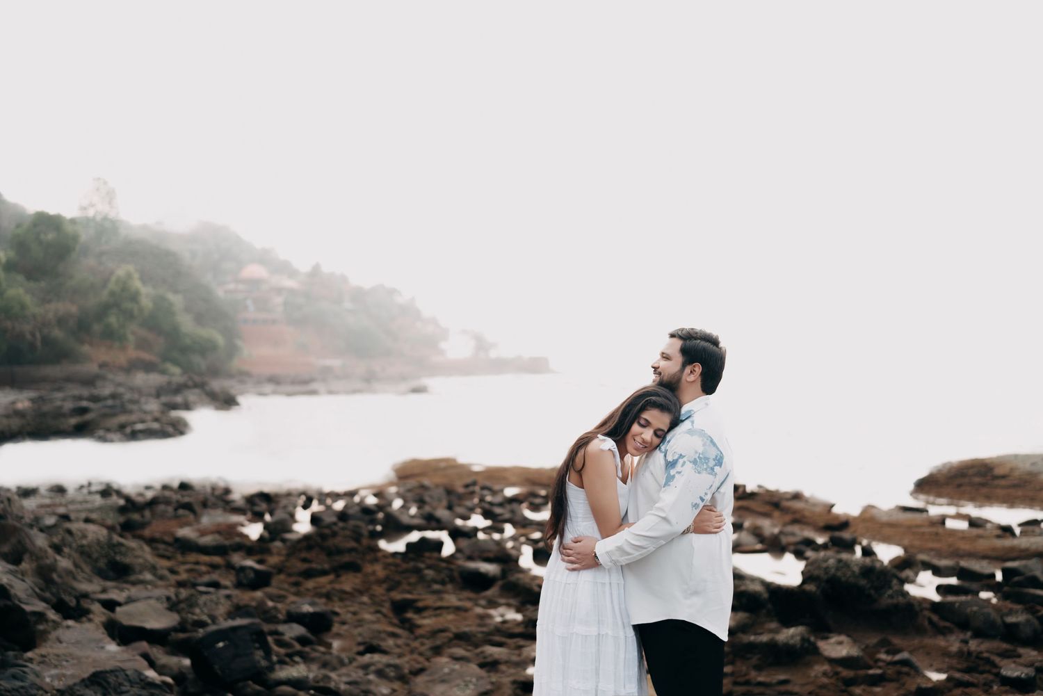 A couple embraces on a rocky beach shoreline with misty coastal cliffs in the background.
