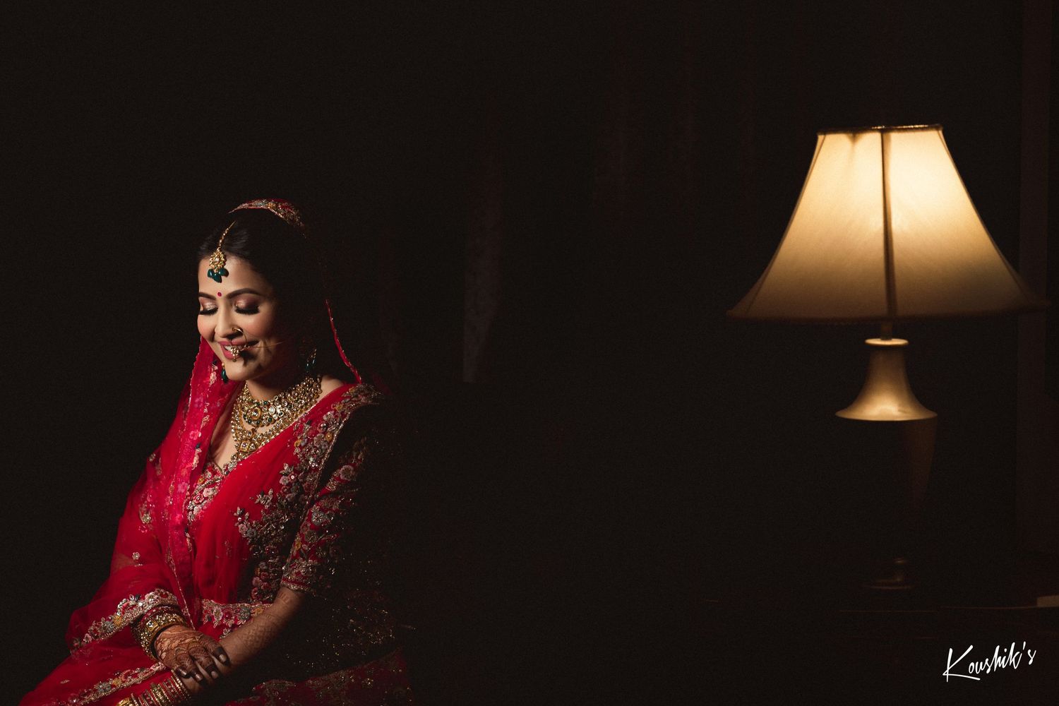 A dramatic low-light scene with a glowing table lamp and traditional red bridal attire visible in the shadows.