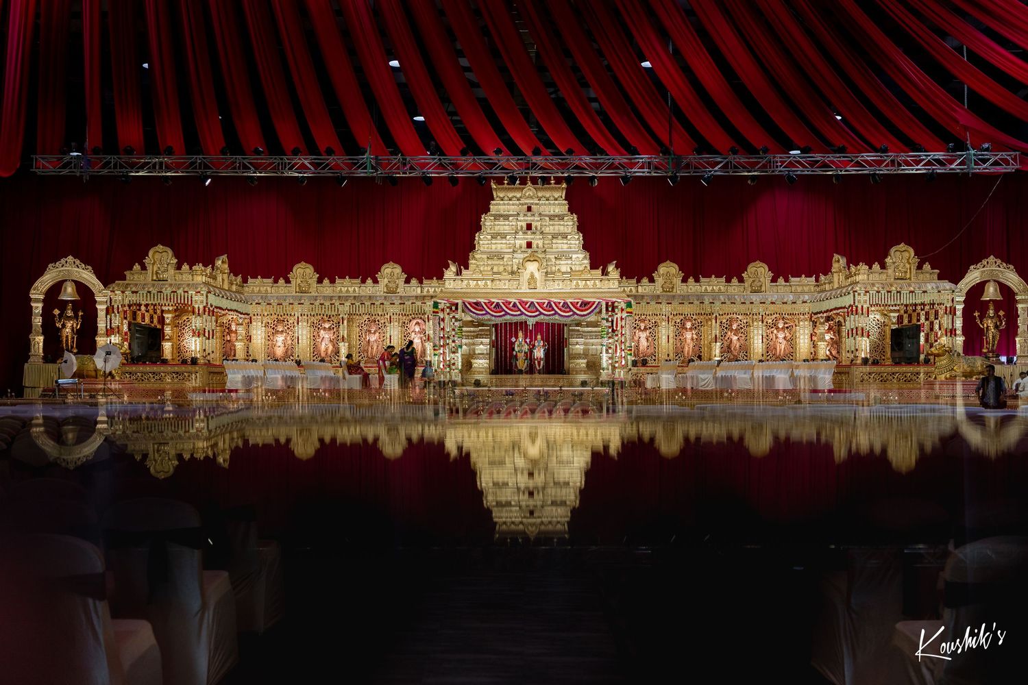 Illuminated temple architecture with ornate golden structures reflected in still water against a dark red background.