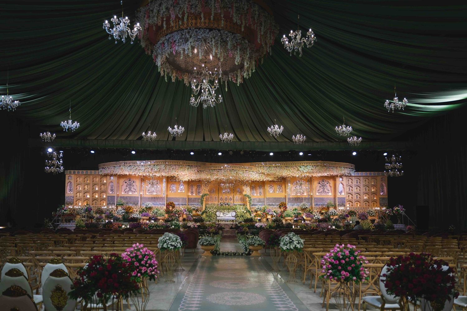 Elegant ballroom reception setup with draped green ceiling, crystal chandeliers and floral arrangements on display tables.