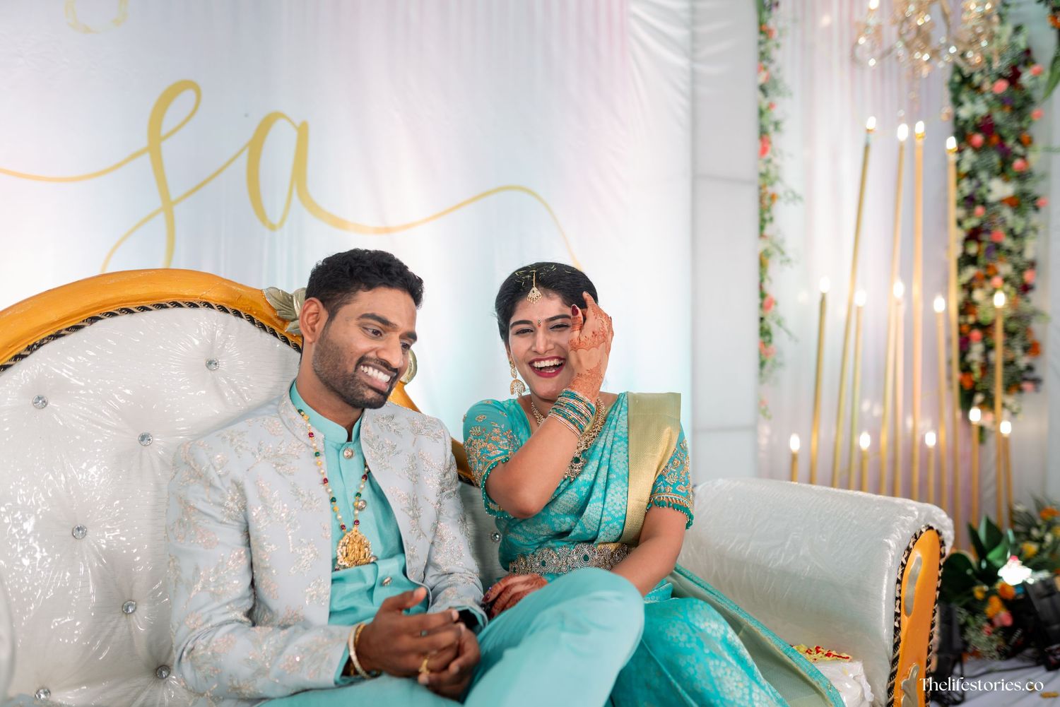 A couple in traditional Indian attire sits together on an elegant white sofa with gold trim against a decorated backdrop.