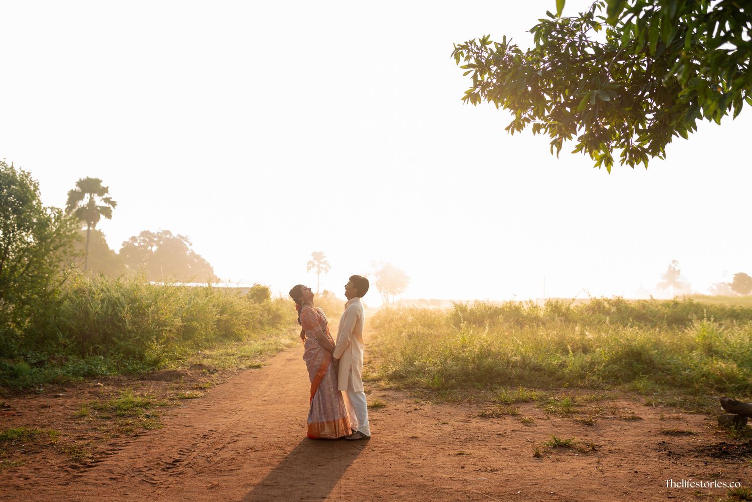 Two people stand together on a dirt path at sunset with golden sunlight streaming through trees in the background.