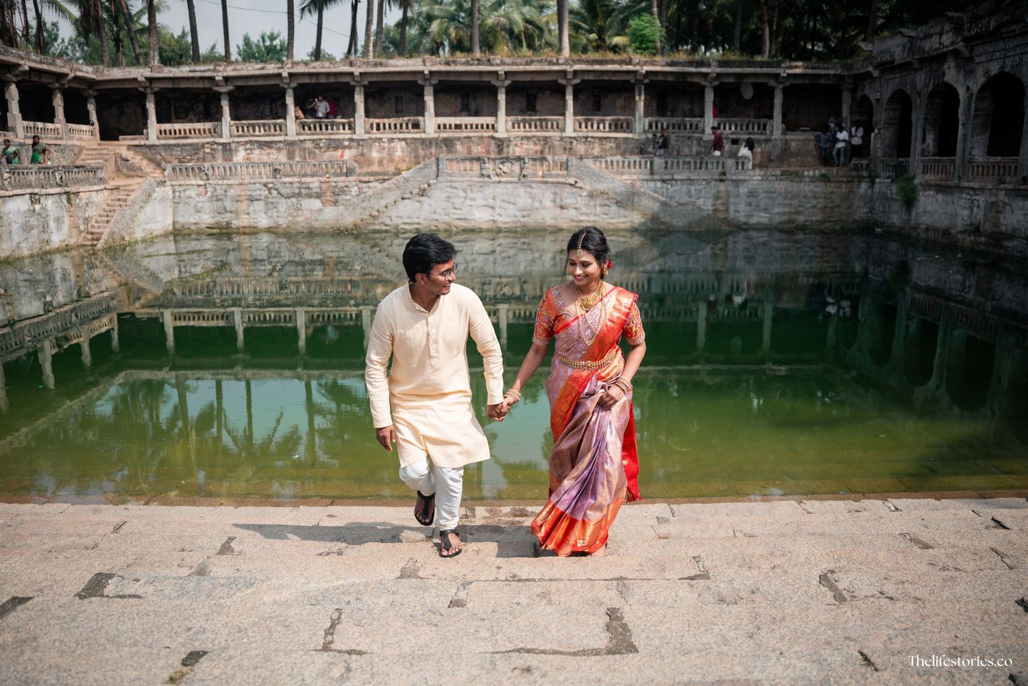 A couple walks together beside an ancient stepwell with green water in India, with traditional architecture visible.