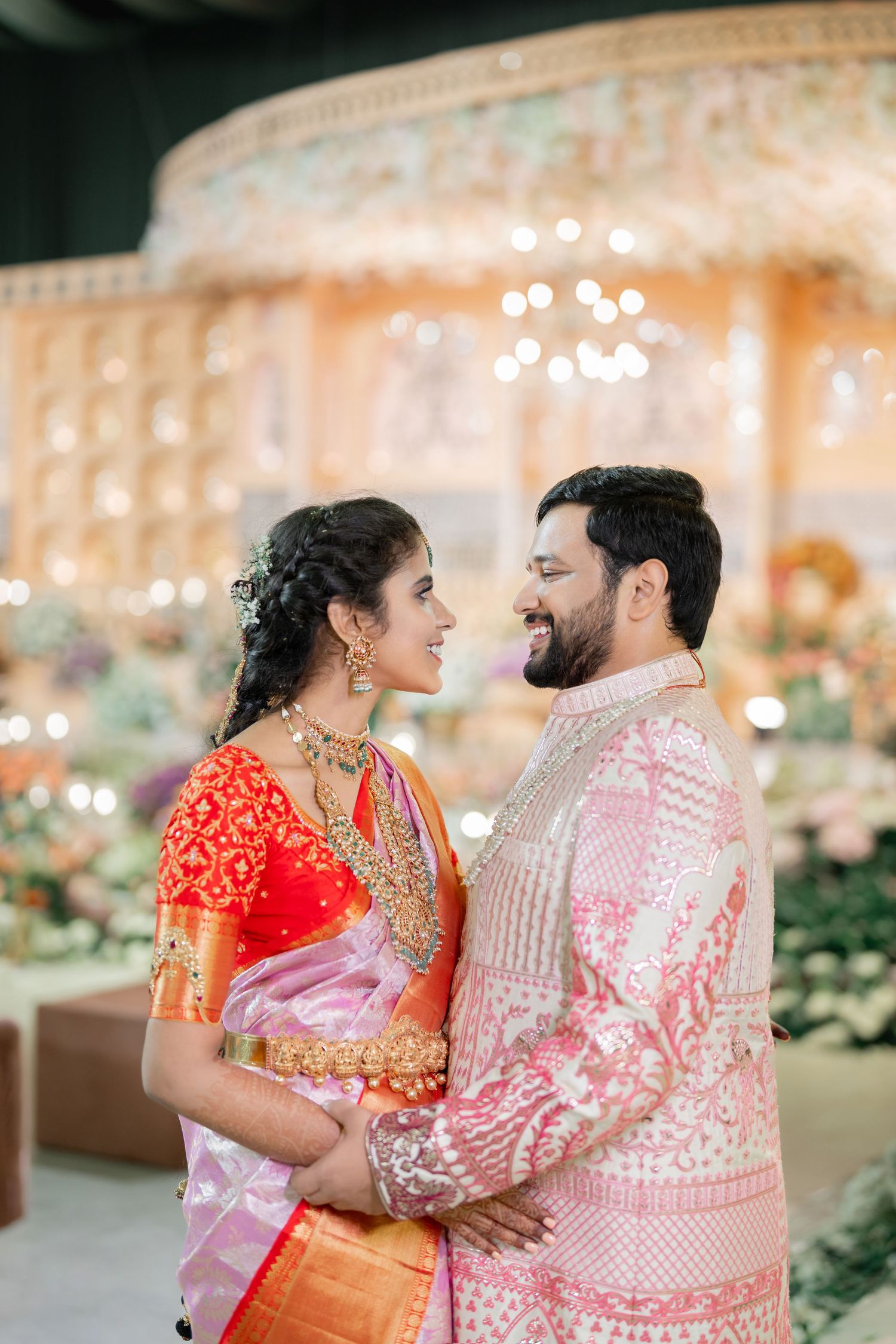 A couple in traditional Indian wedding attire share an intimate moment against a backdrop of twinkling lights and floral decor.