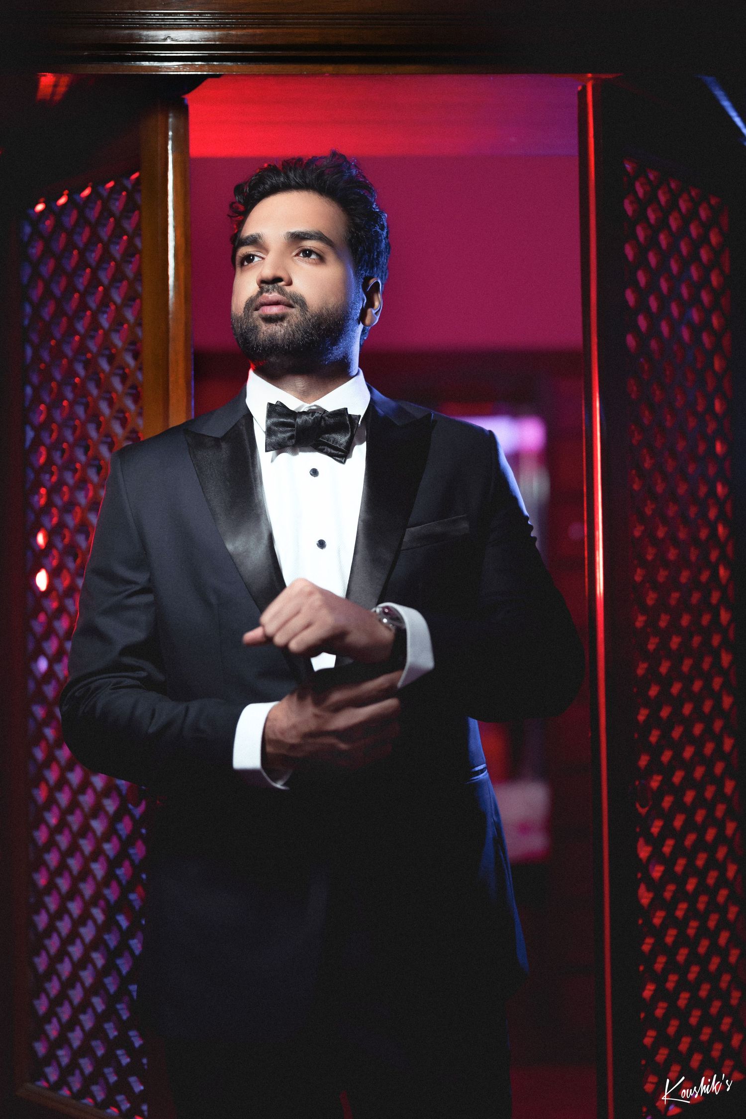 A person in a black tuxedo and bow tie stands confidently against a red-lit backdrop with textured walls.