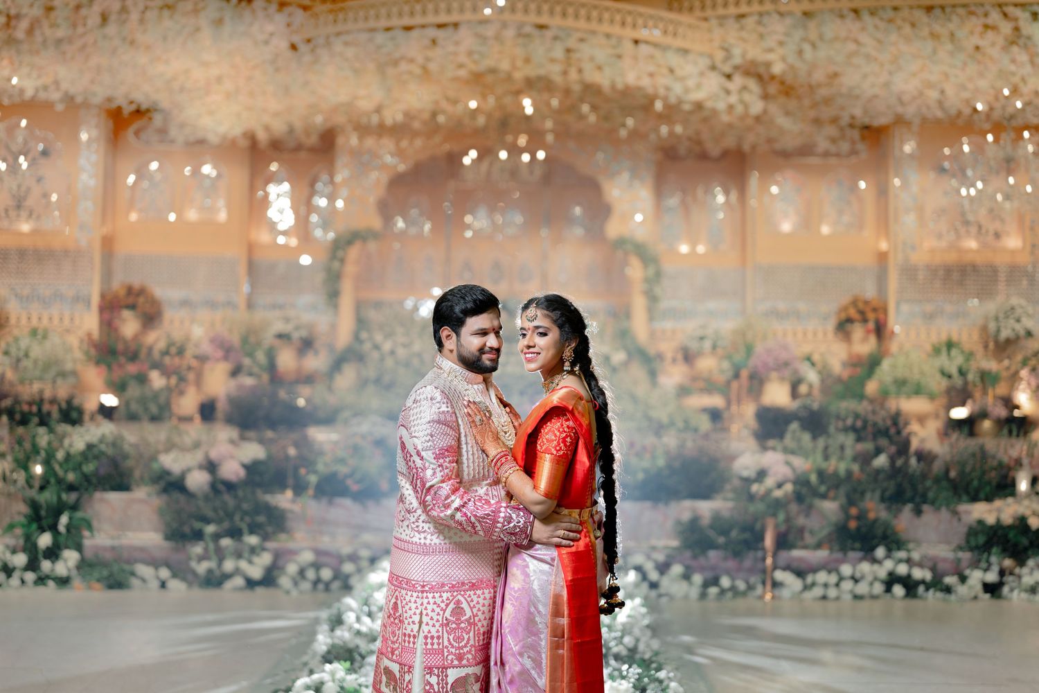 A blurred romantic couple poses in traditional South Asian wedding attire with decorative lights in the background.